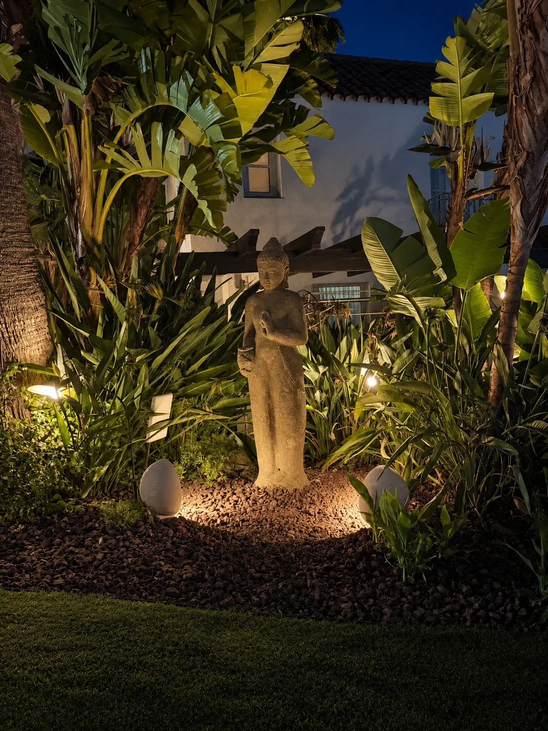 Standing stone sculpture framed by tropical foliage and warm garden lighting at night.